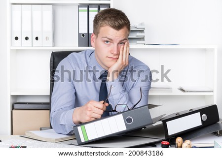 Young thoughtful businessman is sitting in front of many files on his desk in the office. A shelf is in the background. The man is looking to the camera.