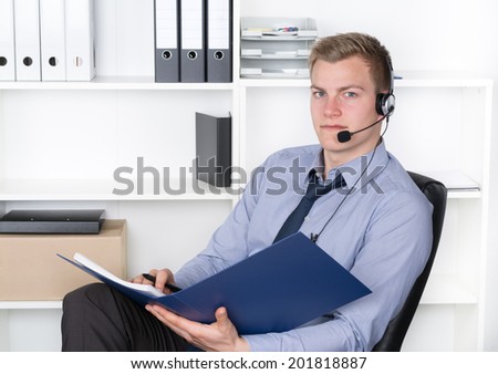 Young relaxed businessman with headset is holding a file while sitting in the office. A shelf is in the background. The man is looking to the camera.