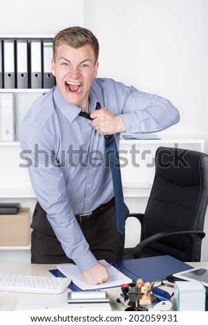 Young furious businessman is tearing open his tie while standing at his desk in the office. A shelf is in the background. The man is looking to the camera.