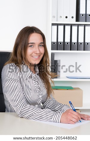 A young smiling businesswoman is writing at a sheet of paper with a blue pen at the desk in the office. A shelf is standing in the background. The Woman is looking to the camera.