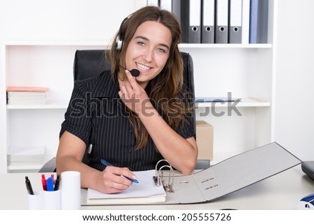 A young smiling businesswoman with headset is writing into a file while sitting at the desk in the office. A shelf is standing in the background. The Woman is looking to the camera.