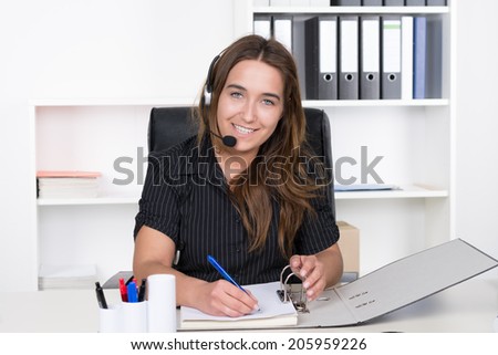 A young smiling businesswoman with headset is writing into a file while sitting at the desk in the office. A shelf is standing in the background. The Woman is looking to the camera.