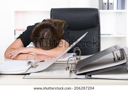 A young frustrated businesswoman is lying in front of a pile of files on the desk in the office. A shelf is standing in the background.