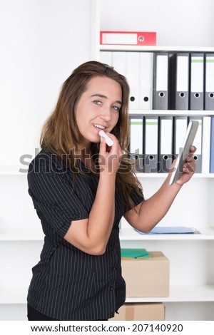 A young businesswoman is using a tablet as make up mirror while standing in the office. A shelf is standing in the background. The woman is looking to the camera.