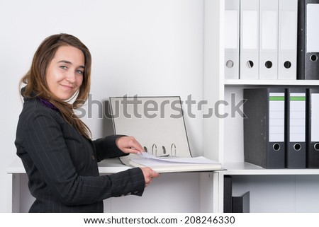 A young smiling businesswoman is browsing a file in front of a shelf in the office. The woman is looking to the camera.