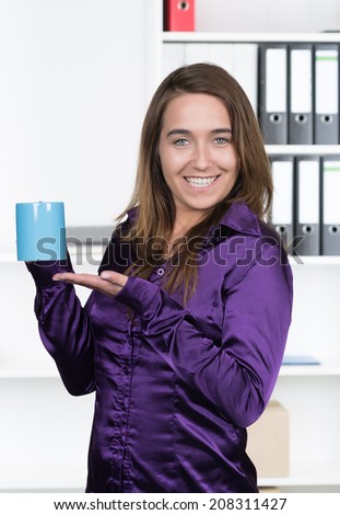 A young smiling businesswoman is presenting a blue cup while standing in the office. A shelf is standing in the background. The woman is looking to the camera.