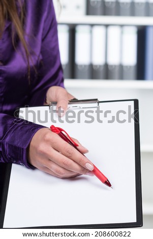 A businesswoman is pointing to a clipboard with a red pen while standing in the office. A shelf is standing in the background.