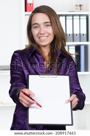 A young smiling businesswoman is pointing to a clipboard with a pen while standing in the office. A shelf is standing in the background. The woman is looking to the camera.