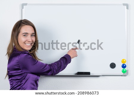 A young smiling businesswoman is pointing to a whiteboard with a pen while standing in the office. The woman is looking to the camera.