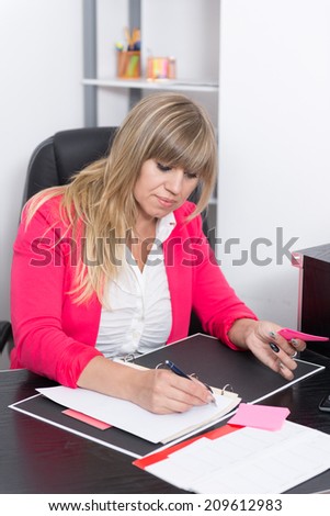 Woman is writing into a file while sitting at the desk in the office. The woman is looking to the file.