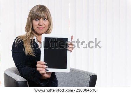 A businesswoman is showing a tablet while sitting on a chair. The woman is looking to the camera. Face is in focus, tablet is blurred.