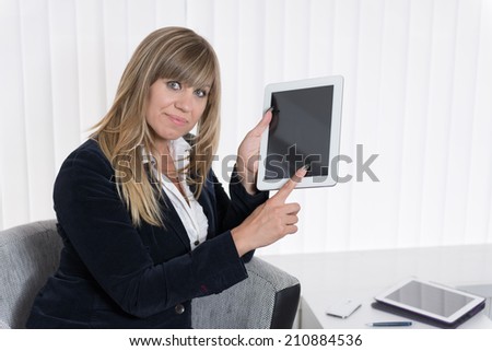 A businesswoman is pointing to a tablet with her finger while sitting on a chair. The woman is looking to the camera.