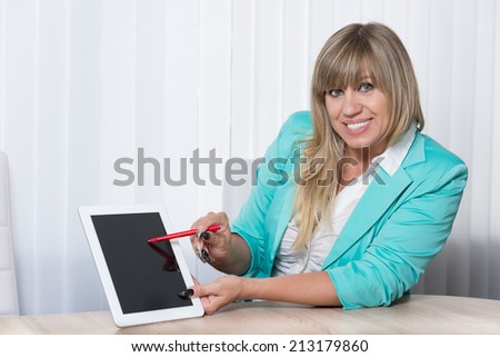 Smiling businesswoman is pointing with a pen to a tablet while sitting at a table in the office. The woman is looking to the camera.