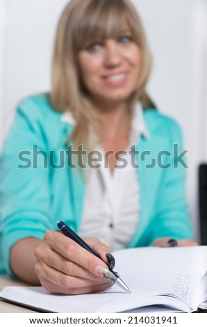 A businesswoman is writing into an appointment calendar which is lying in front of her while she is sitting at a table in the office. The pen is in focus, face is blurred.