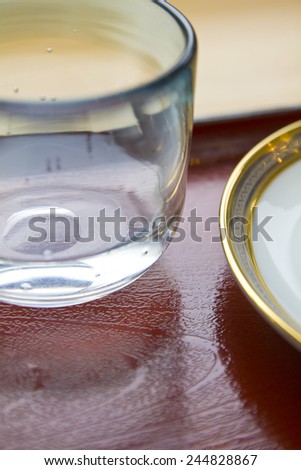 Close up shot of an empty water glass and a white and gold dish on a red wood table.