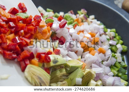 Pouring red peppers, artichokes, peas and other Mediterranean vegetables on a pan in a kitchen.