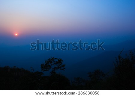 The blue sky at sunset.Mountain on background,tree are black shape.Sun nearly go down.