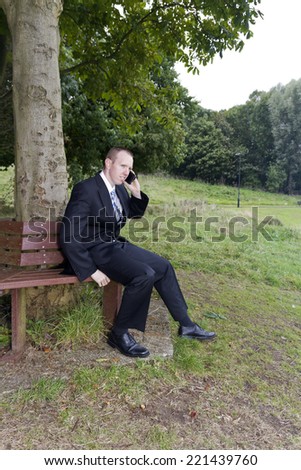 Young man, business man, dressed in suit. Sitting on a park bench, talking on a mobile telephone or cellphone.