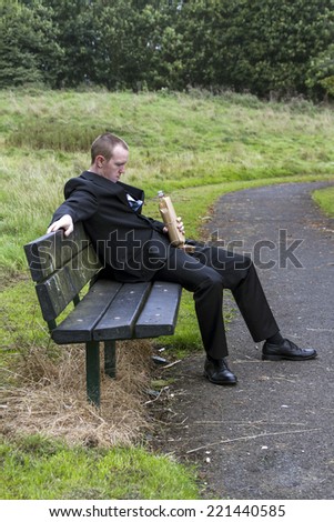 Young man, business man, dressed in suit. Sitting on park bench drinking from bottle wrapped in brown paper.