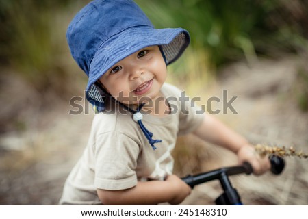 Cute 2 year old mixed race Asian Caucasian boy wearing a blue hat rides his balance bike outside in the summer sun