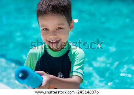 Cute mixed race Asian Caucasian boy happily plays with a water gun in a backyard swimming pool in the summer sun