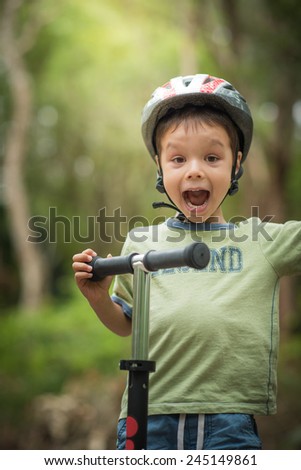 Cute 4 year old mixed race Asian Caucasian boy wearing a bike helmet cheerfully plays on his scooter on a green forest road