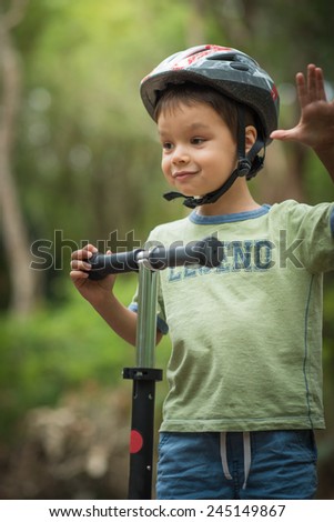 Cute 4 year old mixed race Asian Caucasian boy wearing a bike helmet cheerfully plays on his scooter on a green forest road