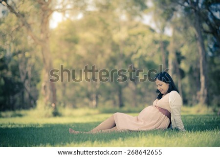 Asian woman 9 months pregnant sitting and relaxing peacefully outside in the grass. Muted tones, faded filtered effects