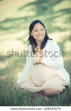 Asian woman 9 months pregnant sitting outside in the grass holding freshly picked dandelions picked by her boys. Looking happily off camera. Muted tones, soft faded filter effects.
