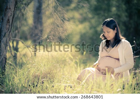 Asian woman 9 months pregnant sitting outside in long grass. Tranquil and natural environment. Muted tones, soft faded filtered effects.