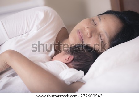 Asian mother sleeps peacefully on her bed with her mixed race 10 day old newborn baby boy in her bedroom