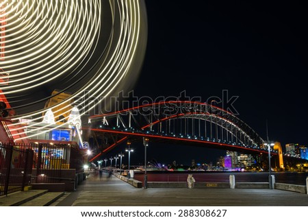 Night view of Sydney Harbour Bridge with Luna Park ferris wheel in foreground and Sydney CBD lights in the background.