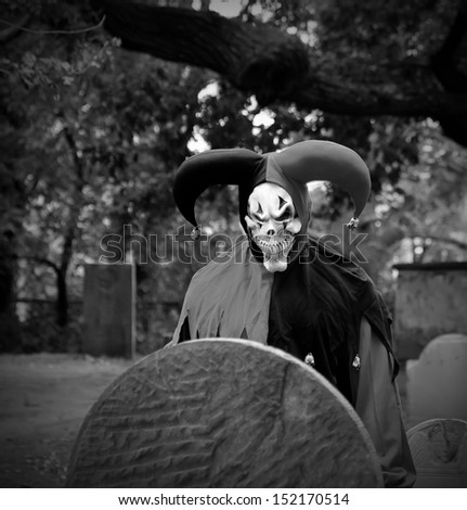 Scary Joker with skeleton face looking at the headstone on the graveyard. Black and White.