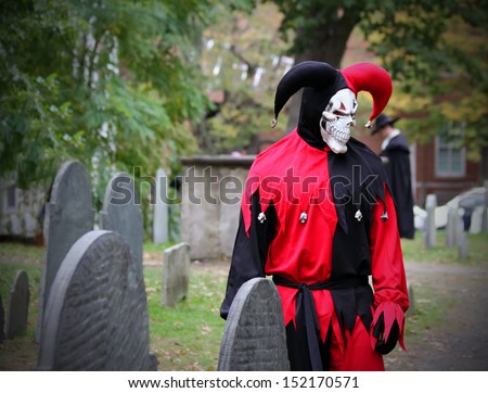 Scary Joker with skeleton face looking at the headstone on the graveyard