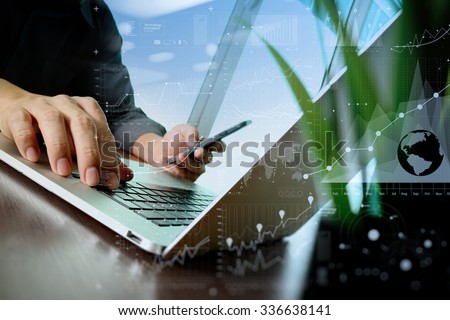 designer hand working and smart phone and laptop and digital business layers with green plant foreground on wooden desk in office