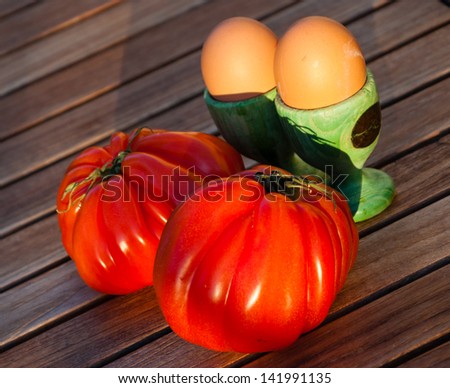 Two shiny ripe beefsteak tomato and two eggs in green wooden egg cups isolated on wooden background. Golden light and long evening shadows.
