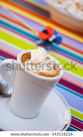 Cup of coffee with whipped cream and funny decorative bug on colorful striped tablecloth.  Reflection on cup and on saucer. Selective focus.