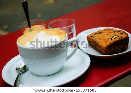 Cup of  cappuccino, slice of carrot pie with walnuts, glass with brown sugar and glass of water on the red table in cafe. Outdoor. Selective focus.