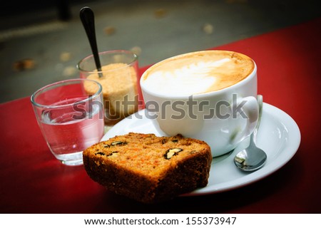Cup of  cappuccino, slice of carrot pie with walnuts, glass with brown sugar and glass of water on the red table in cafe. Outdoor. Selective focus. Shadowed angles.