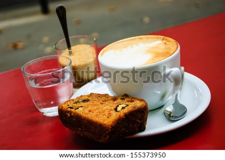 Cup of  cappuccino, slice of carrot pie with walnuts, glass with brown sugar and glass of water on the red table in cafe. Outdoor. Selective focus.