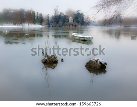 Snow covered boat in the lake and Love Temple at backgrounds.  Lake Daumesnil (Vincennes forest, Paris, France). Soft focus and bleached angles.