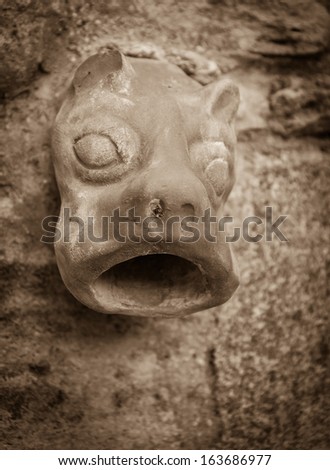 Old ceramic downspout in shape of beast head against stone wall. Closeup. Sepia.