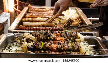 Assortment of  kebabs and grilled sausages and the hands of the seller which prepares the hot dog sandwich at Christmas market in Paris. Selective focus on the upper kebabs.