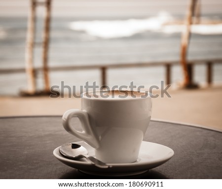 Coffee cup on round table in cafe terrace on the old pier with rusty railing and the view on the sea with waves in cold cloudy day. Loneliness idea. Aged photo.