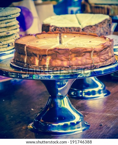 Cheesecake on metal cake stand and other cakes and cookies on grungy wooden bar in rustic coffee shop. Selective focus on the cake crust. Aged photo.