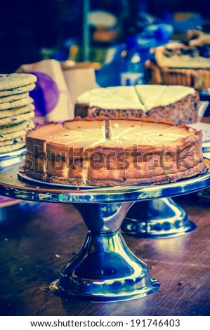 Cheesecake on metal cake stand and other cakes and cookies on grungy wooden bar in rustic coffee shop. Selective focus on the cake crust. Aged photo.