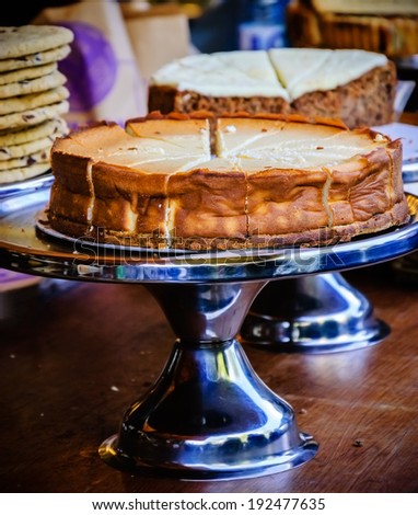 Cheesecake on metal cake stand and other cakes and cookies on grungy wooden bar in rustic coffee shop. Selective focus on the cake crust.