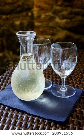 Ornate pitcher with cold white wine and two empty glasses on wicker table in front of rough stone wall in rustic restaurant. Reflection. A game of light and shadow. Selective focus on the pitcher.