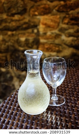 Ornate pitcher with cold white wine and one empty glass on wicker table in front of rough stone wall in rustic restaurant. Reflection. A game of light and shadow. Selective focus on the pitcher.