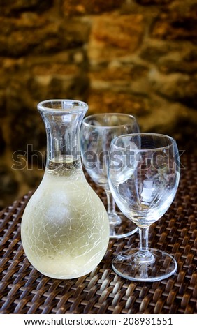 Ornate pitcher with cold white wine and two empty glasses on wicker table in front of rough stone wall in rustic restaurant. Reflection. A game of light and shadow. Selective focus on the pitcher.
